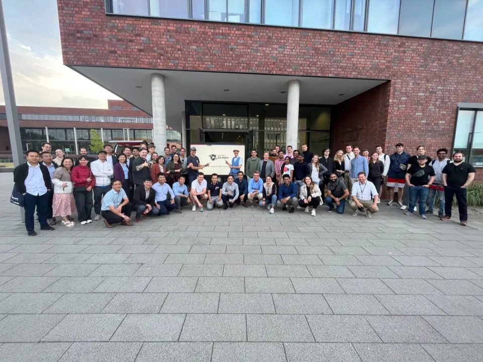 Gruppenfoto vieler Teilnehmender vor einem Backsteingebäude, in der Mitte ein Bildschirm mit dem Himmel-der-Möwen-Logo